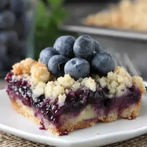 A slice of blueberry pie bar on a white plate with fresh blueberries