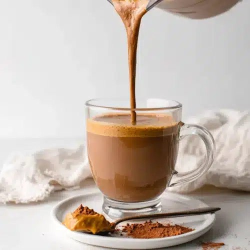 Creamy coffee smoothie being poured into a glass mug with peanut butter and cocoa powder nearby