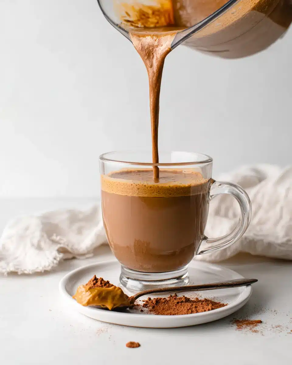 Creamy coffee smoothie being poured into a glass mug with peanut butter and cocoa powder nearby