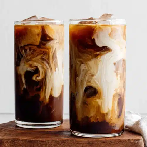Two glasses of cold brew coffee with milk swirls and ice, served on a wooden board