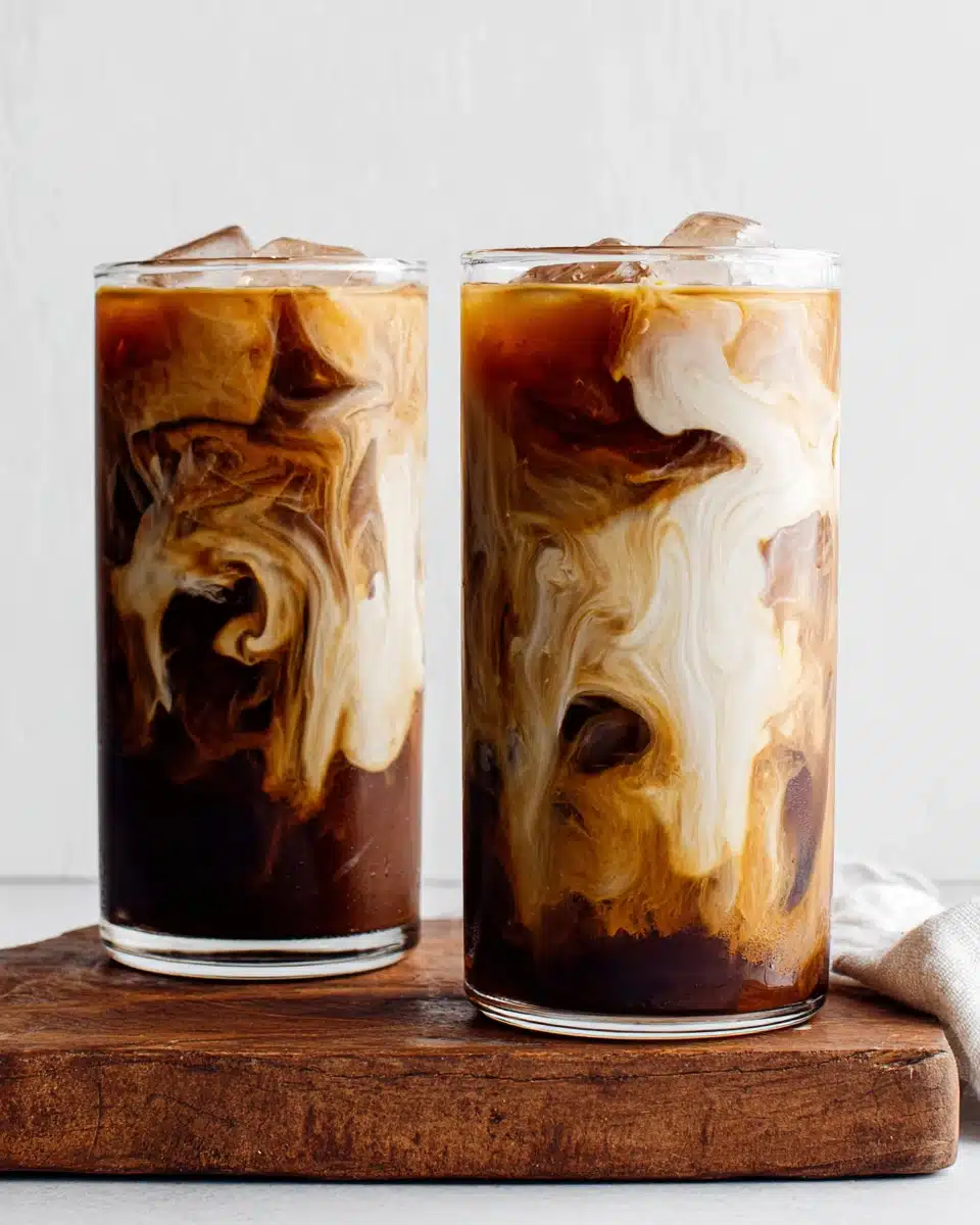 Two glasses of cold brew coffee with milk swirls and ice, served on a wooden board