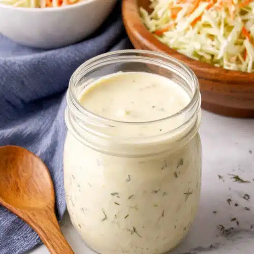 Creamy coleslaw dressing in a glass jar with fresh coleslaw in the background