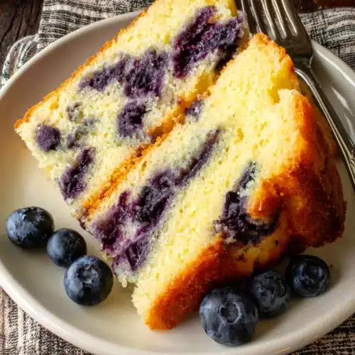 Moist eggless blueberry cake slice with fresh blueberries and fork on white plate