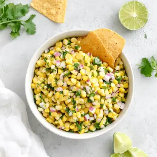 Fresh corn salsa in a white bowl with tortilla chips, lime, and cilantro