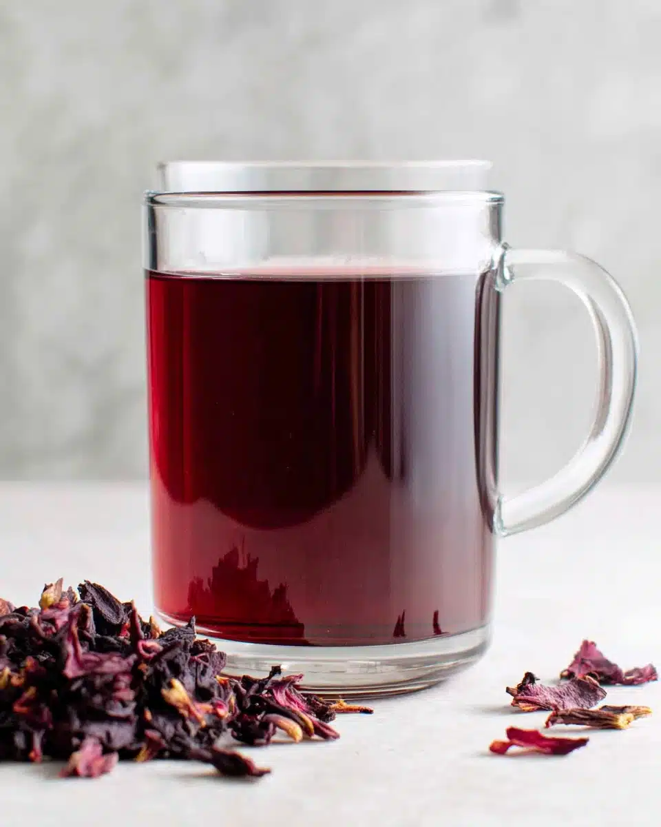A glass mug of hibiscus tea with dried petals nearby