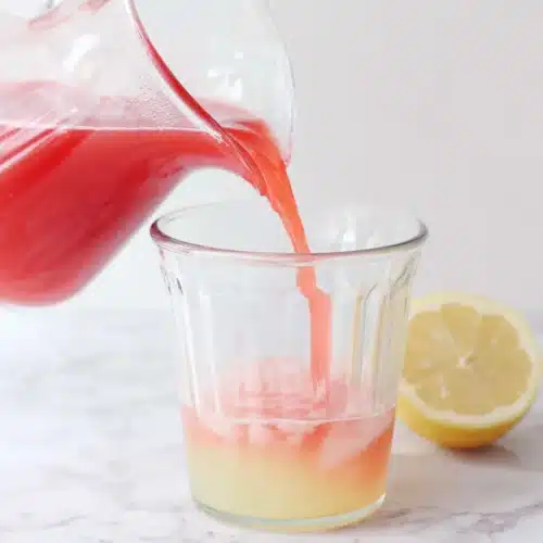 Hand pouring cranberry juice into pink lemonade pitcher with lemon slice nearby