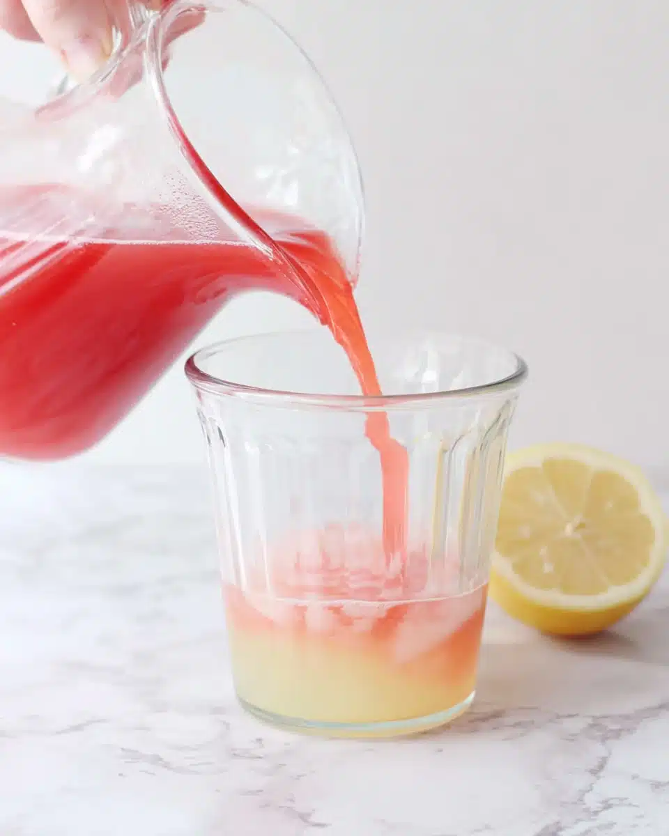 Hand pouring cranberry juice into pink lemonade pitcher with lemon slice nearby