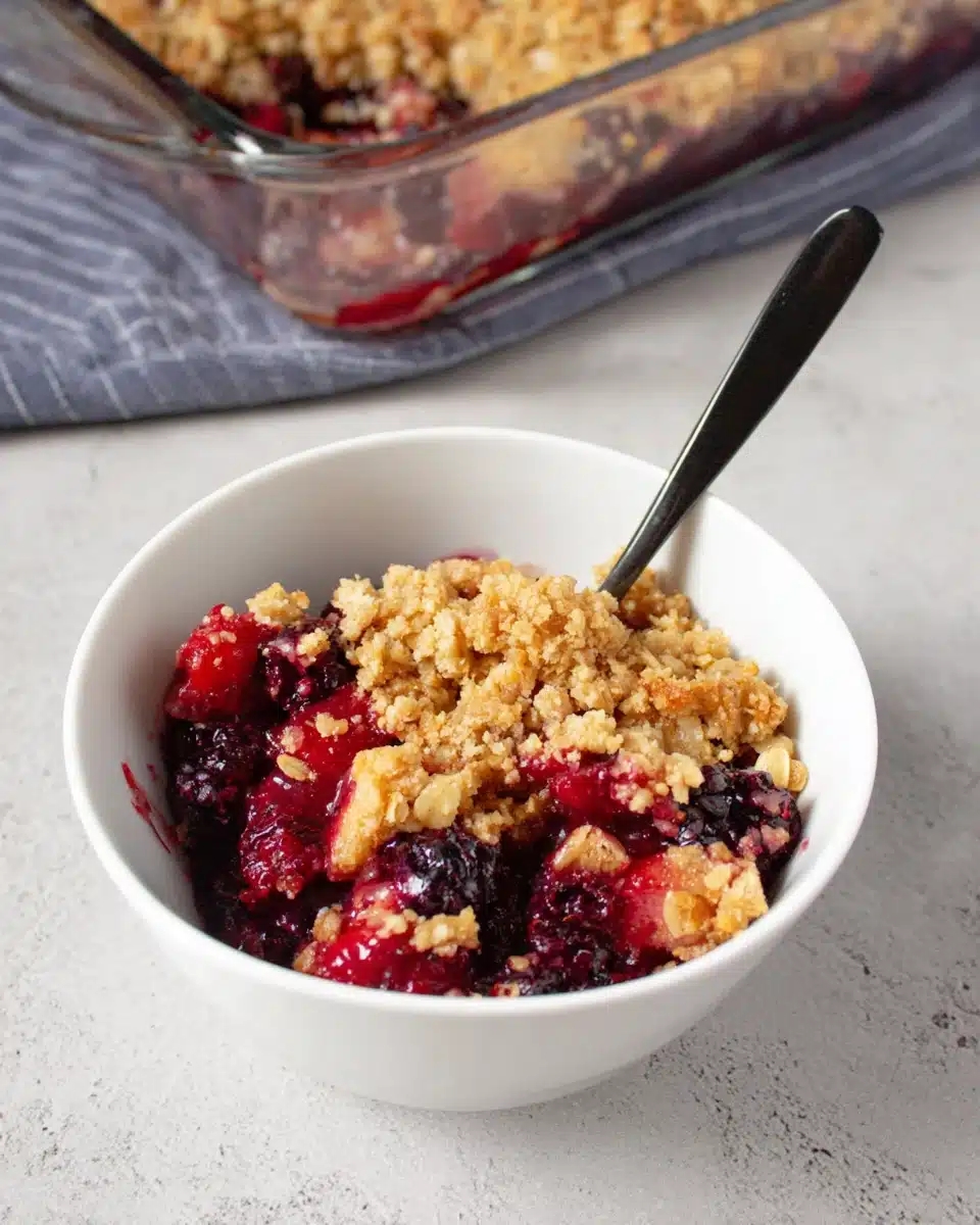 Triple berry crisp in a white bowl with golden oat topping and black spoon