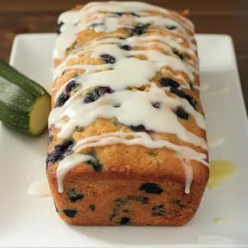 Blueberry zucchini bread with lemon glaze on a white plate, fresh blueberries and zucchini nearby