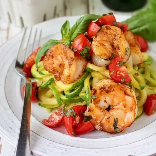 Garlicky tomato-basil shrimp with zoodles on a white plate, garnished with fresh basil and cherry tomatoes