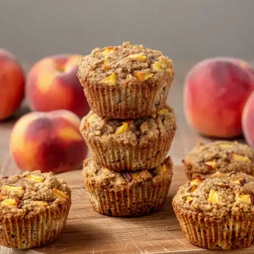 Gluten-free peach muffins stacked with fresh peaches in background