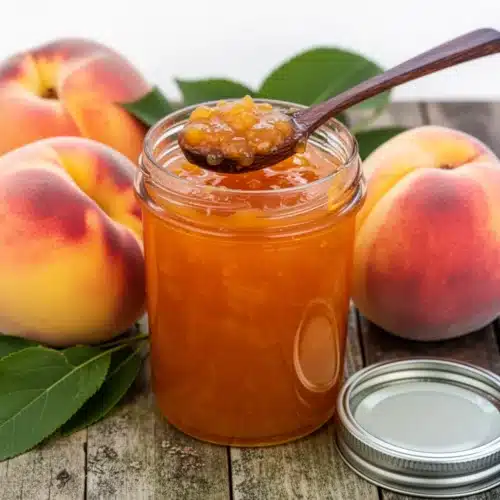 Low sugar peach jam in a glass jar with fresh peaches and green leaves on a wooden table