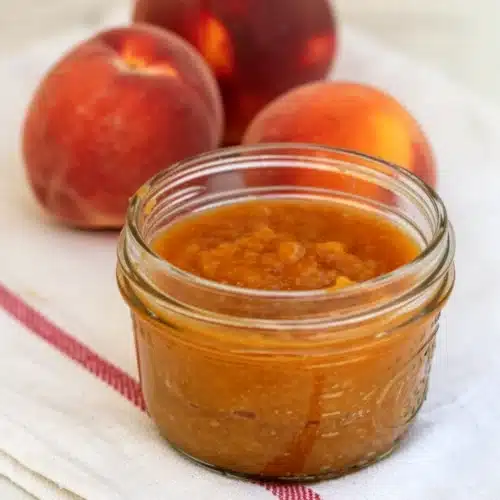 Old-fashioned peach butter in a glass jar with fresh peaches on a striped cloth