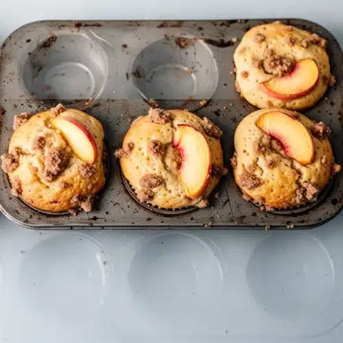 Peach muffins with brown sugar streusel in a muffin tin, topped with fresh peach slices